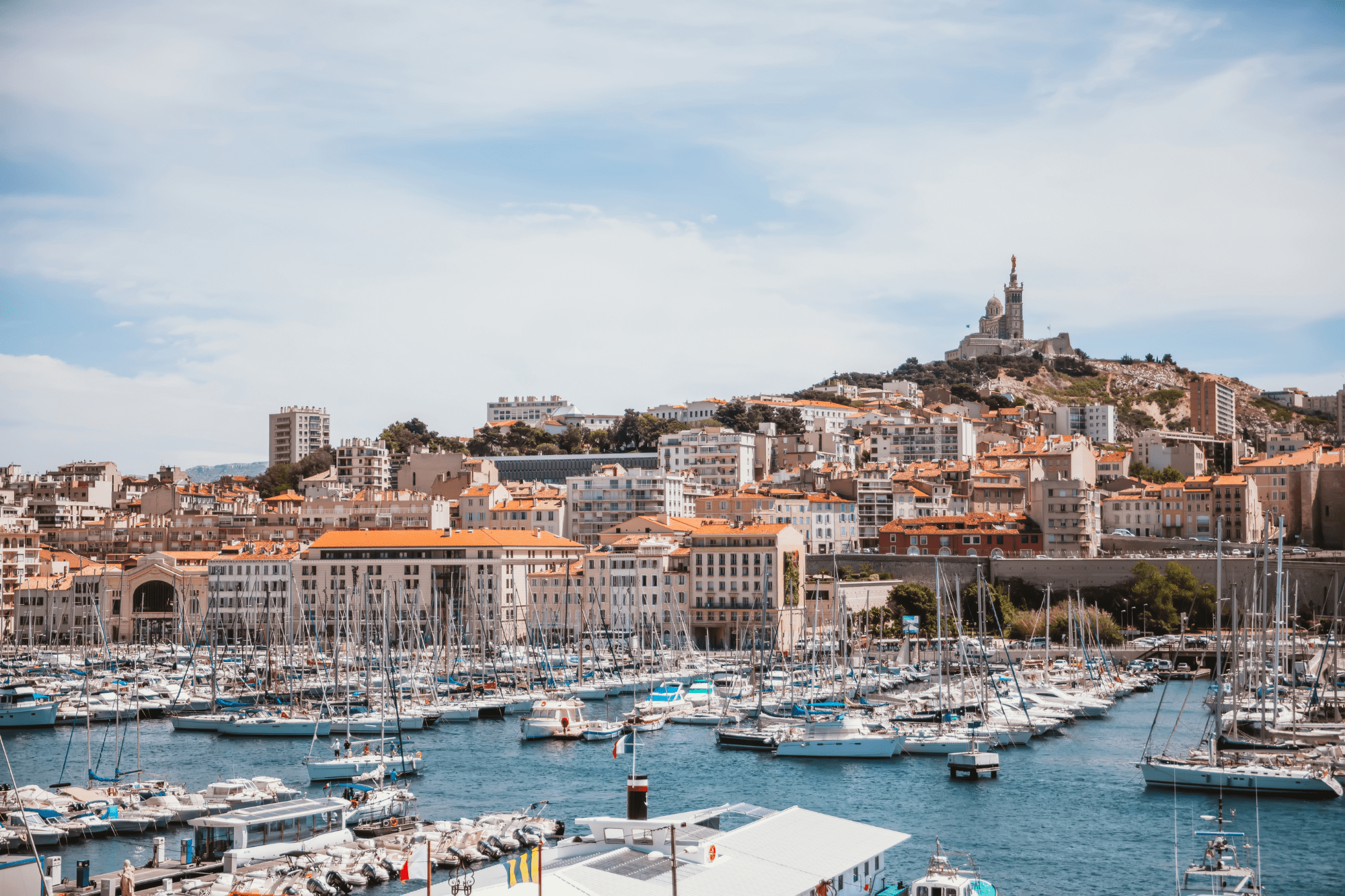 Un port de plaisance rempli de voiliers amarrés dans un paysage urbain aux toits de tuiles rouges, avec une grande église au sommet d'une colline sous un ciel partiellement nuageux - une scène inspirante pour tout projet d'Agence web Marseille ou de création de site internet Marseille.
